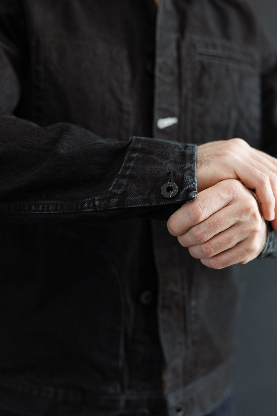 Close-up of front left sleeve cuff and button details on men's black denim jacket, model is male, 5'9
