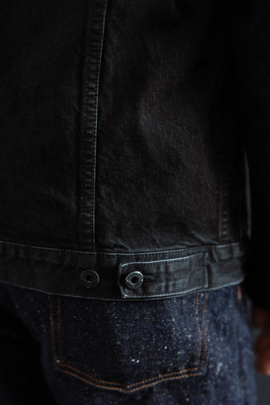 Close-up of men's black denim jacket back hem with button details on male model, rear view