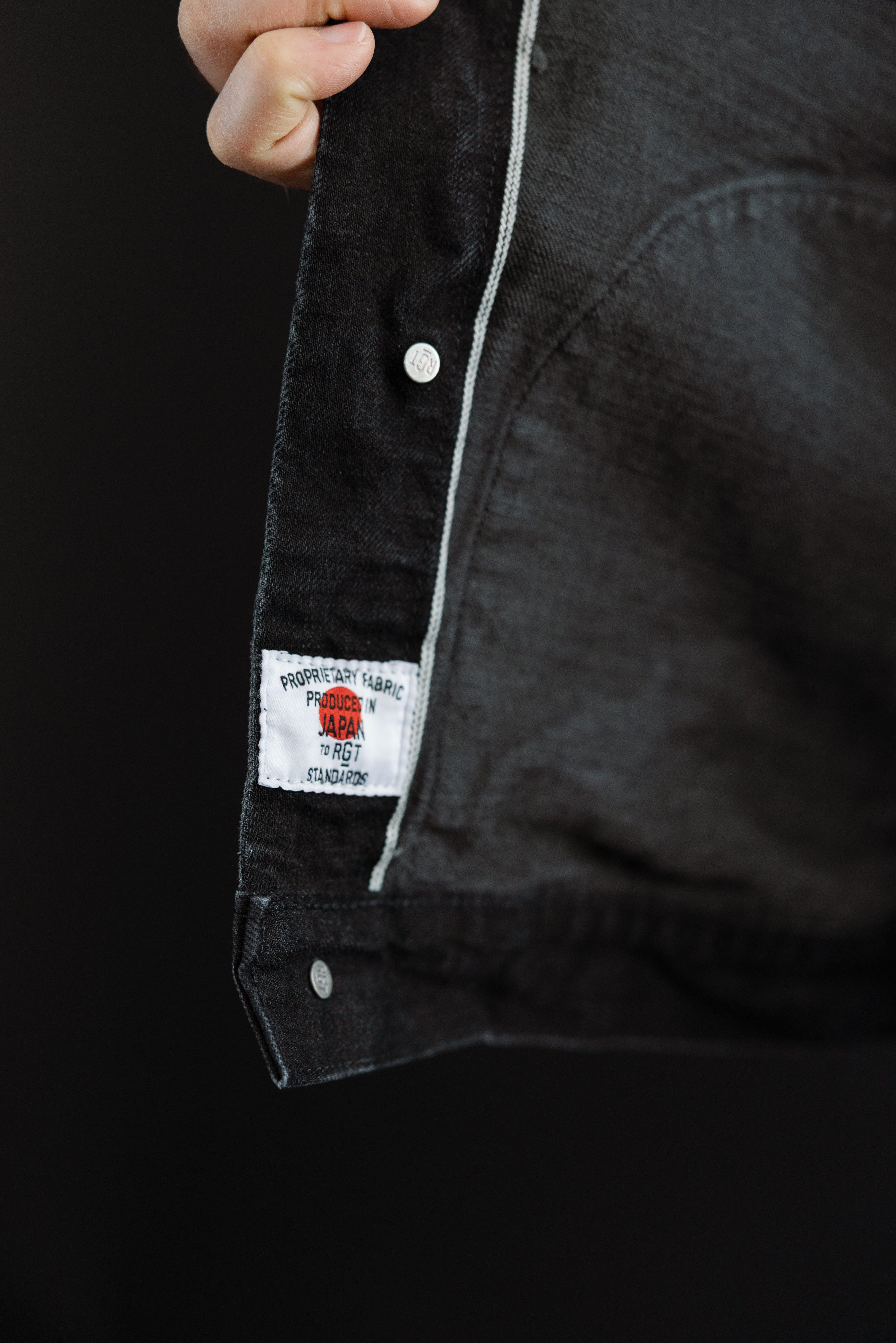 Close-up of inside front hem of black denim jacket, showing selvedge seam, branded rivet, and fabric tag.