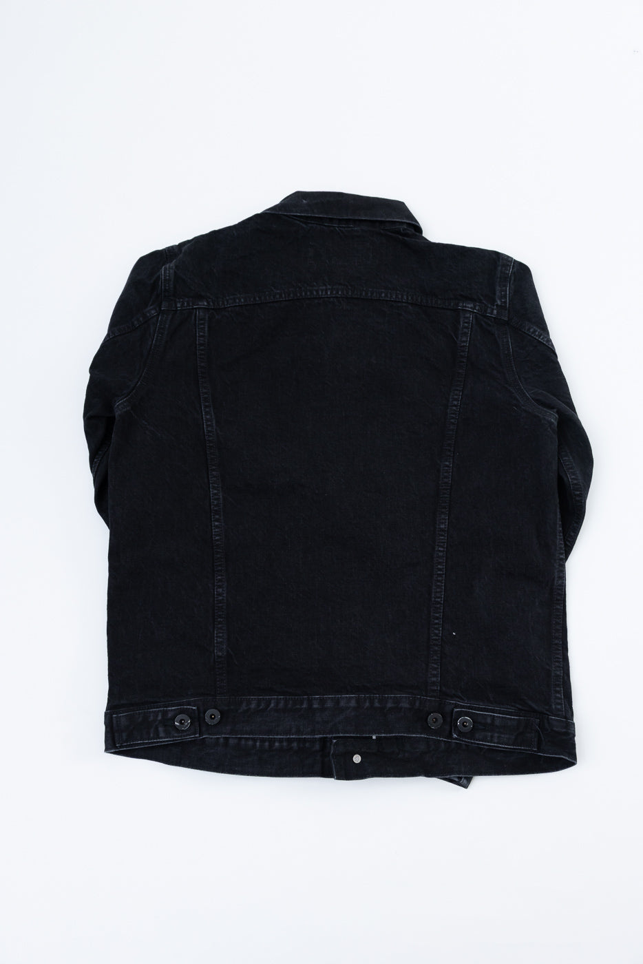 Flat lay of black denim jacket, back view, showing buttoned hem and panel seam details
