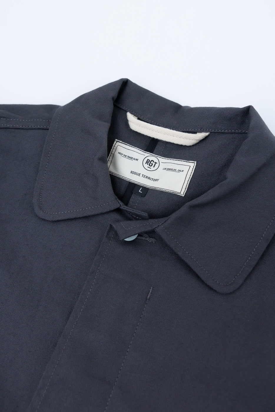 Close-up of front collar and tag on grey dry waxed canvas trench coat, showing texture and stitching detail.