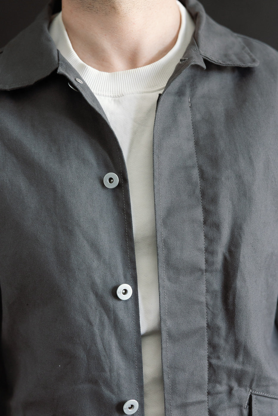 Close-up front view of grey dry waxed canvas trench coat on male model, highlighting buttons, collar, and fabric texture. Model is 5'9