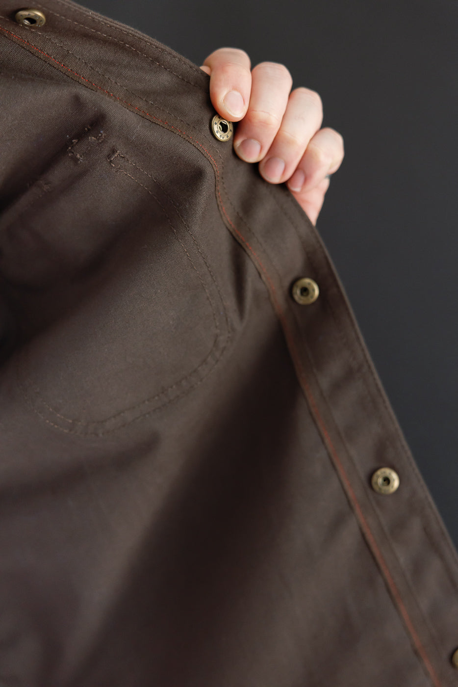 Close-up of a male model's hand holding the front placket of a brown jacket with metal buttons.