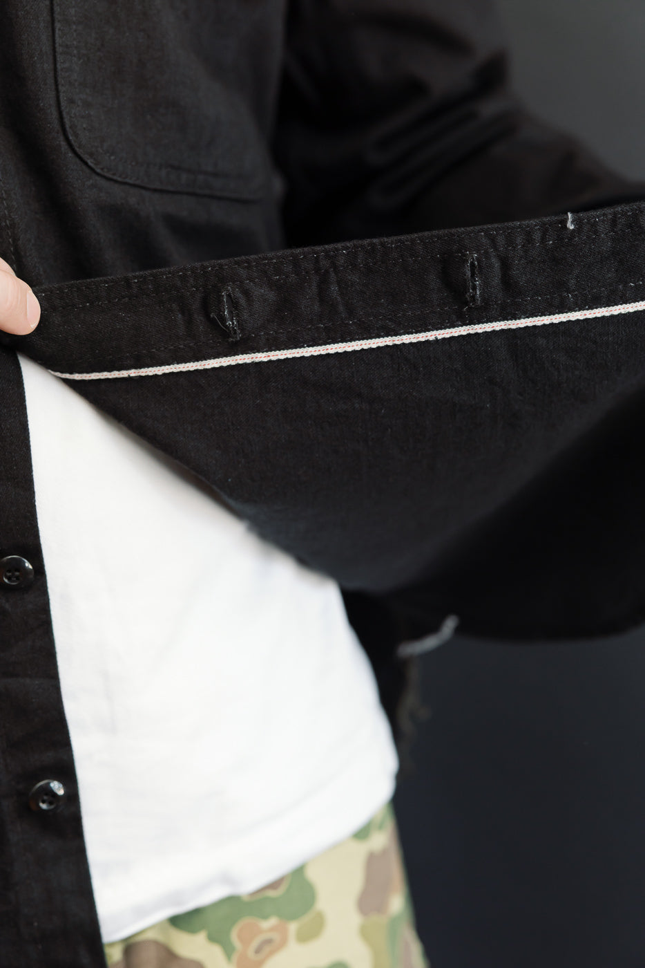 Close-up of black shirt hem with contrast stitching, worn by male model, showing front detail.