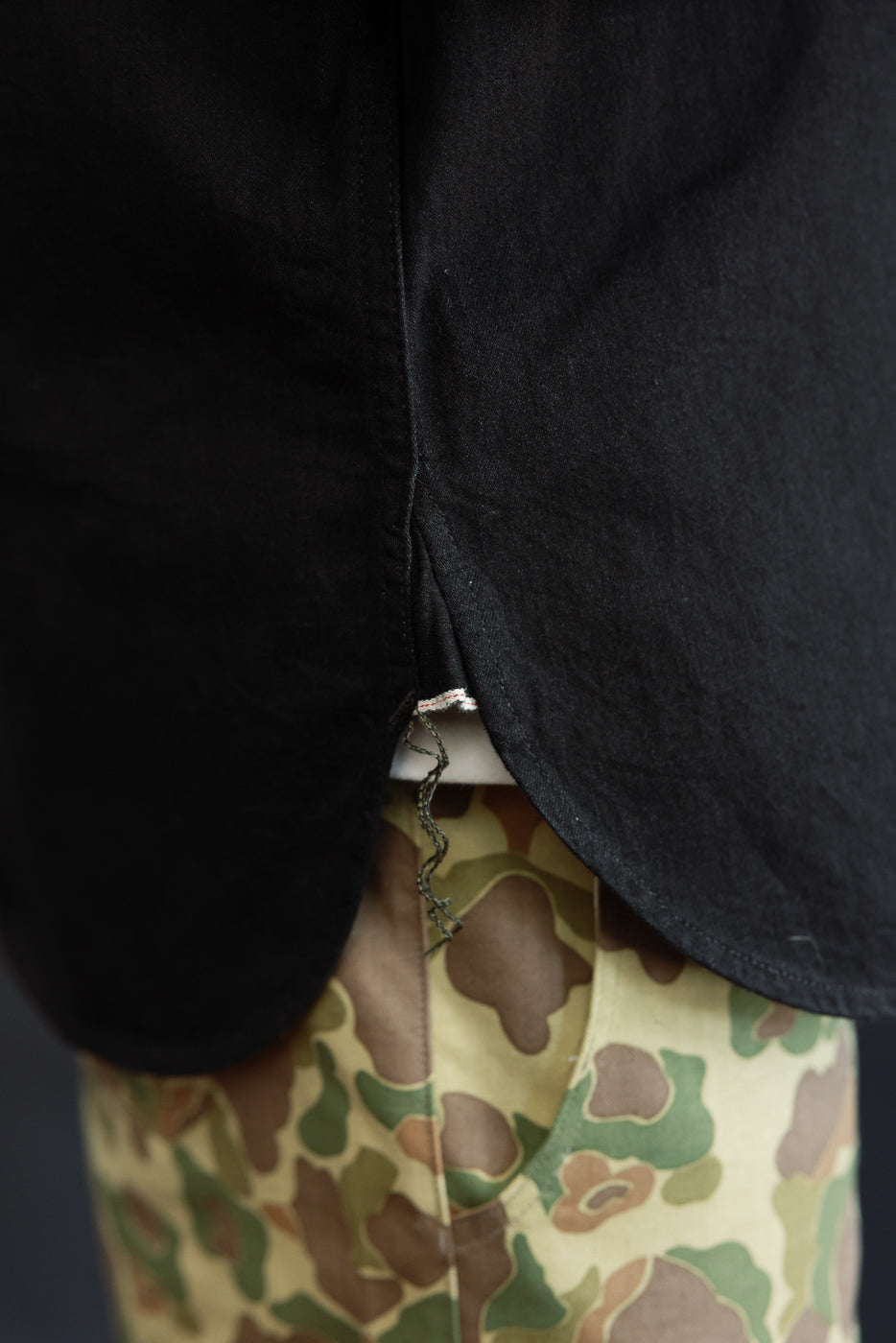 Close-up of black shirt hem with frayed tag, worn by male model over camo pants, back view