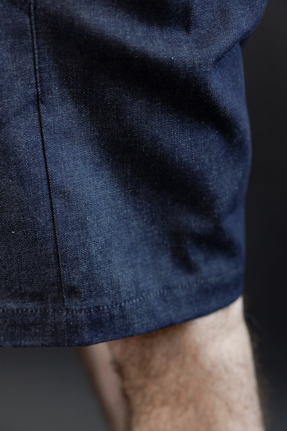 Close-up side view of dark denim shorts hem, worn by male model, showing fabric texture and stitching detail