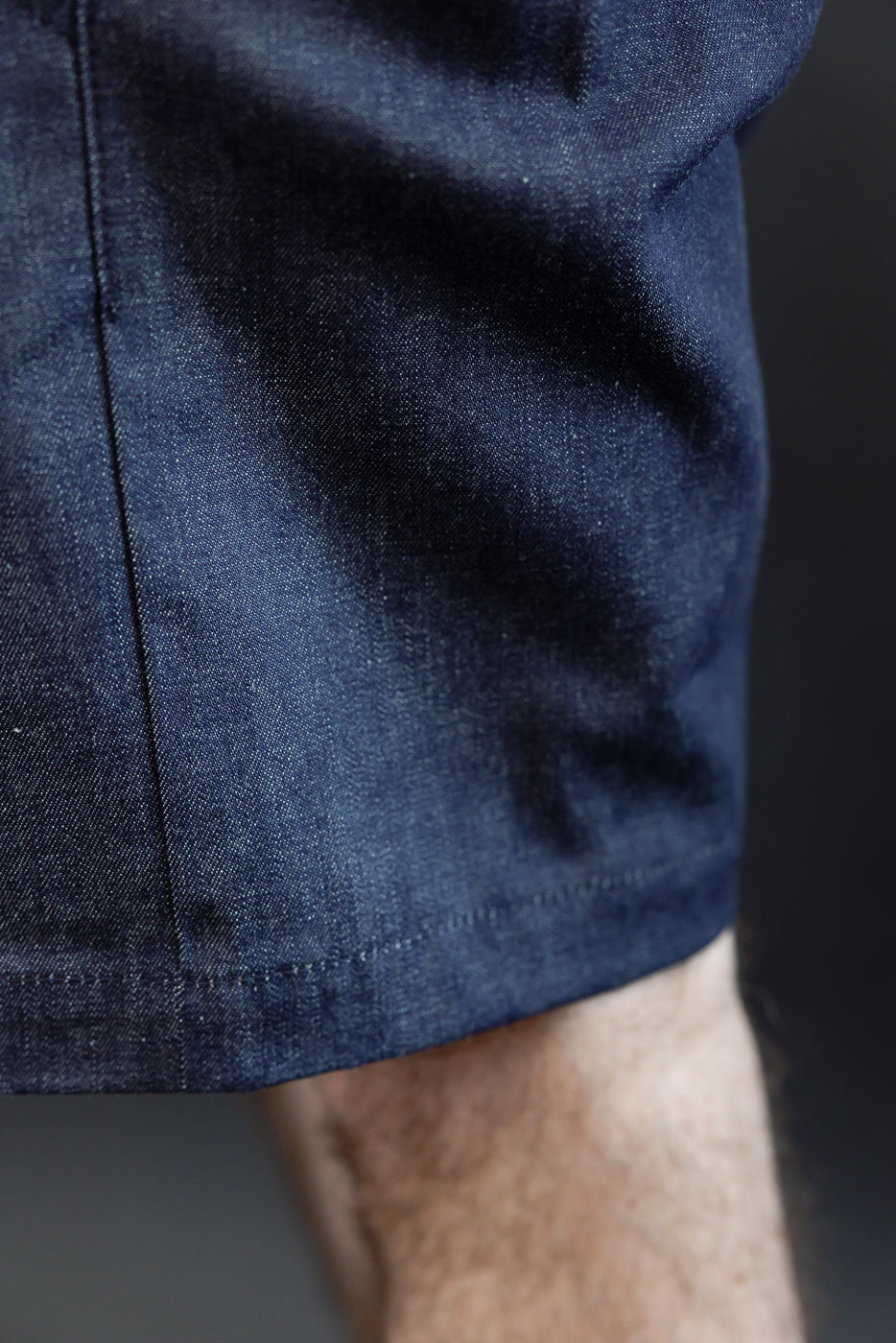 Close-up of dark denim shorts hem, showcasing fabric texture and stitching, worn by male model.