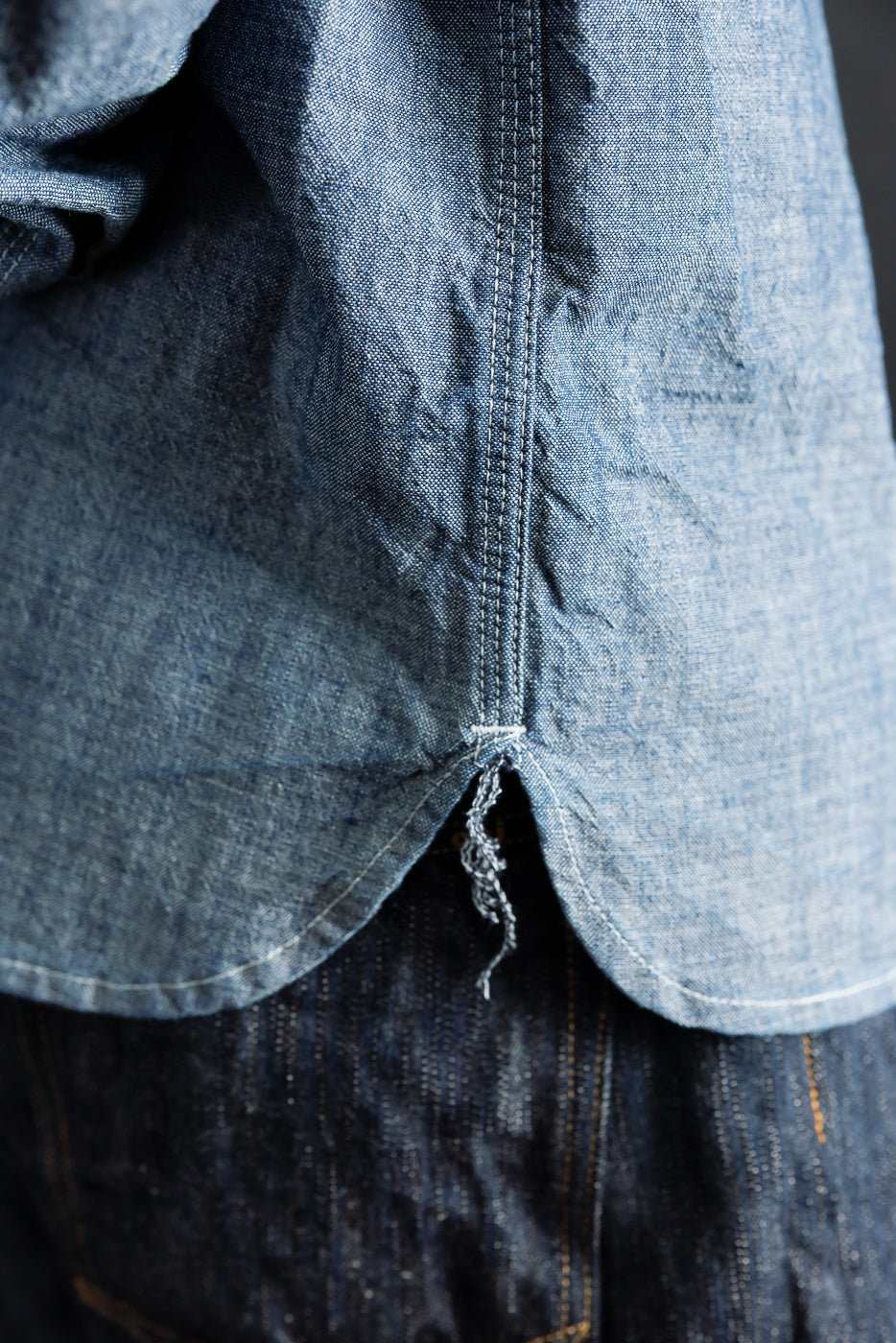 Close-up of side hem detail on rinsed indigo canvas work shirt, showing curved edge stitching and fabric texture.