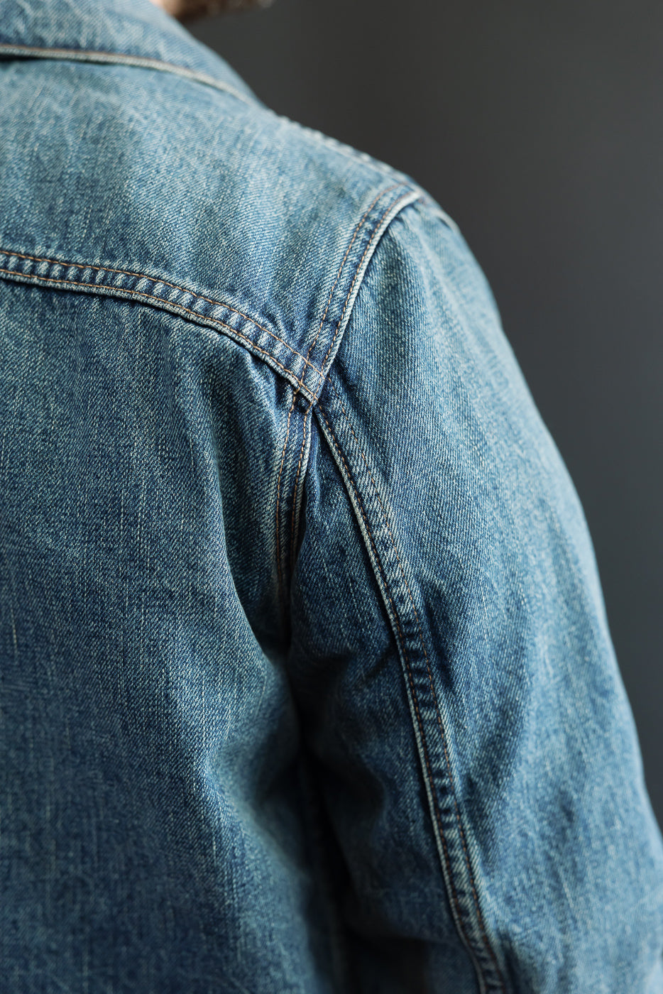 Close-up of male model's left shoulder showing denim jacket's back yoke, stitching, and fabric texture.