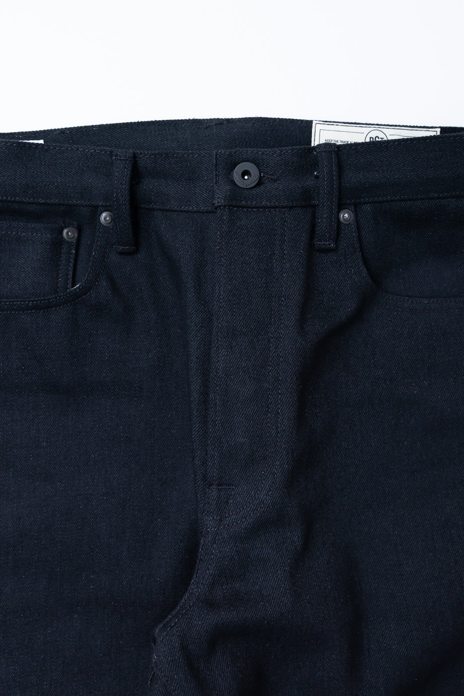 Close-up front view of dark selvedge denim jeans showing waistband, button, and pocket detail.