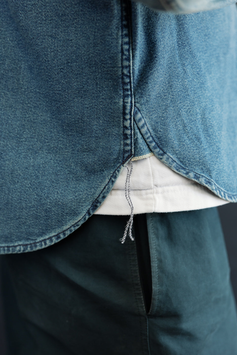Close-up of male model's front hem on denim shirt, showing curved hemline, raw edge detail, and layered white tee underneath.