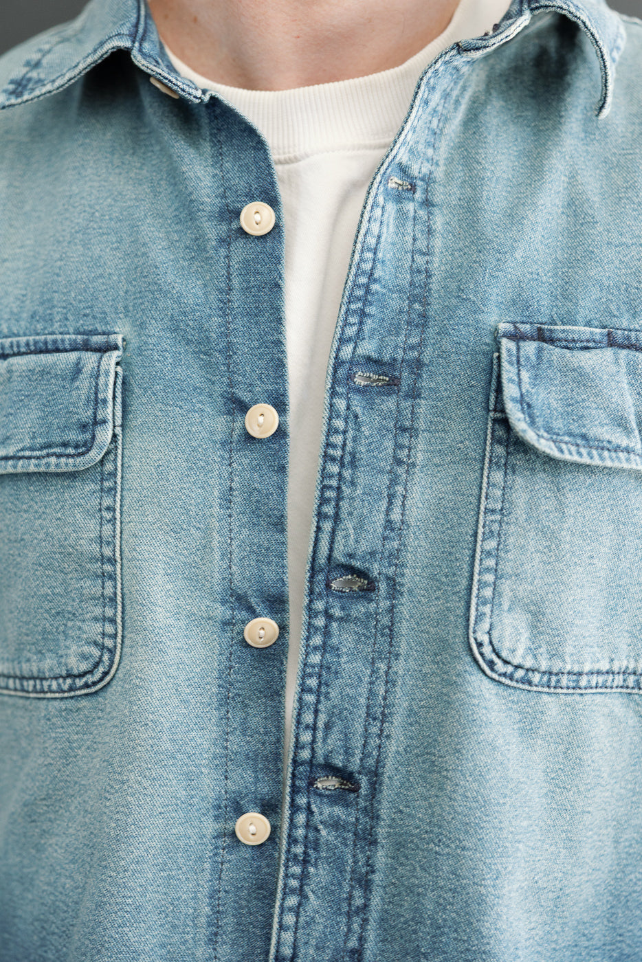Close-up front view of male model wearing light wash denim shirt with button-front, chest pockets, and collar detail.