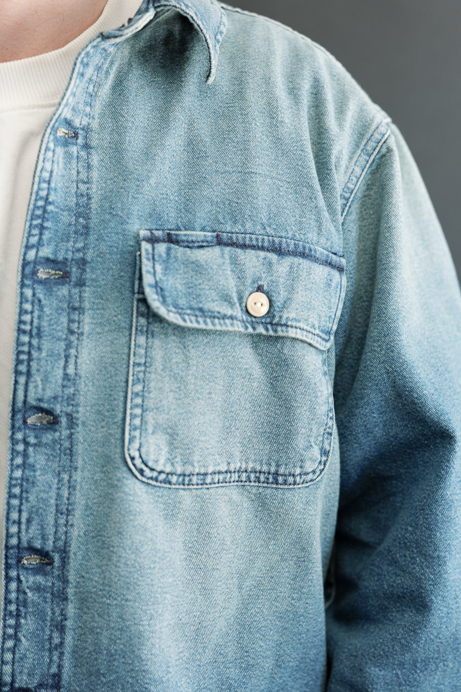 Close-up front view of male model's light wash denim shirt showing chest pocket, button, and fabric texture