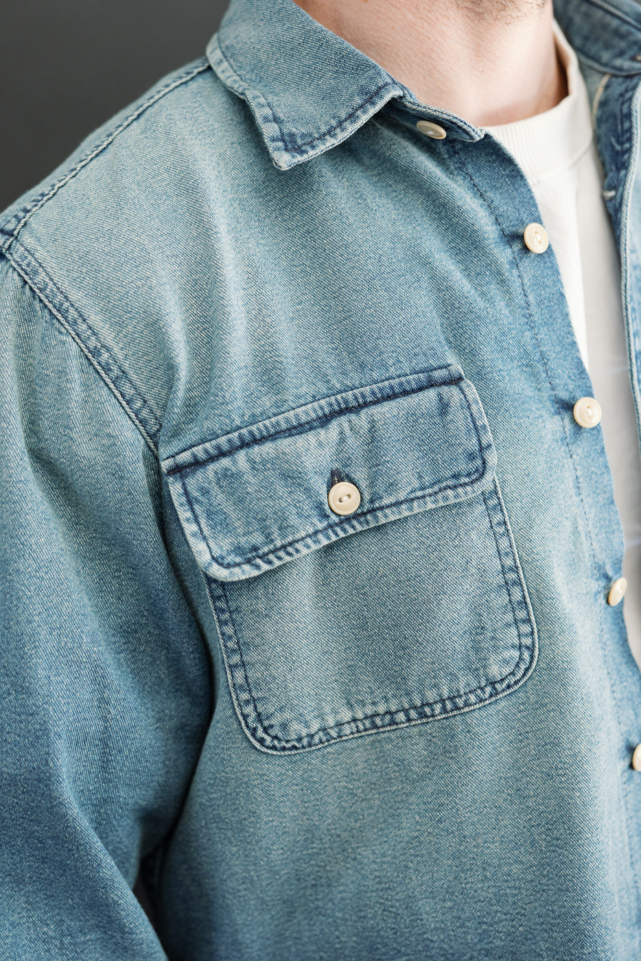 Close-up front view of male model wearing light wash denim shirt with chest pocket, button closure, and collar detail