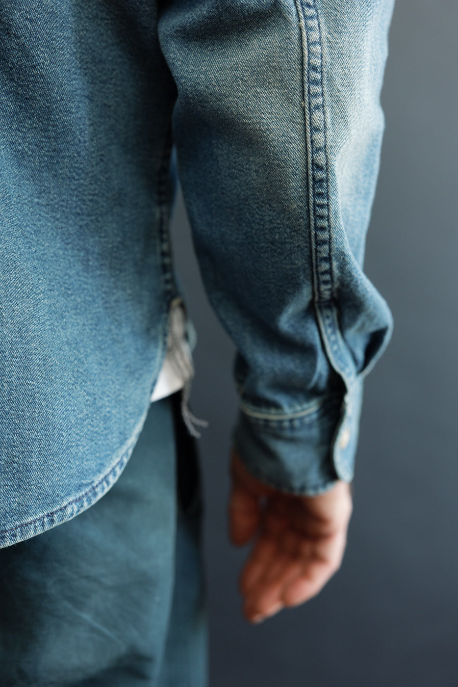 Close-up side view of male model's denim shirt sleeve, showing button cuff, selvedge stitching, and fabric texture