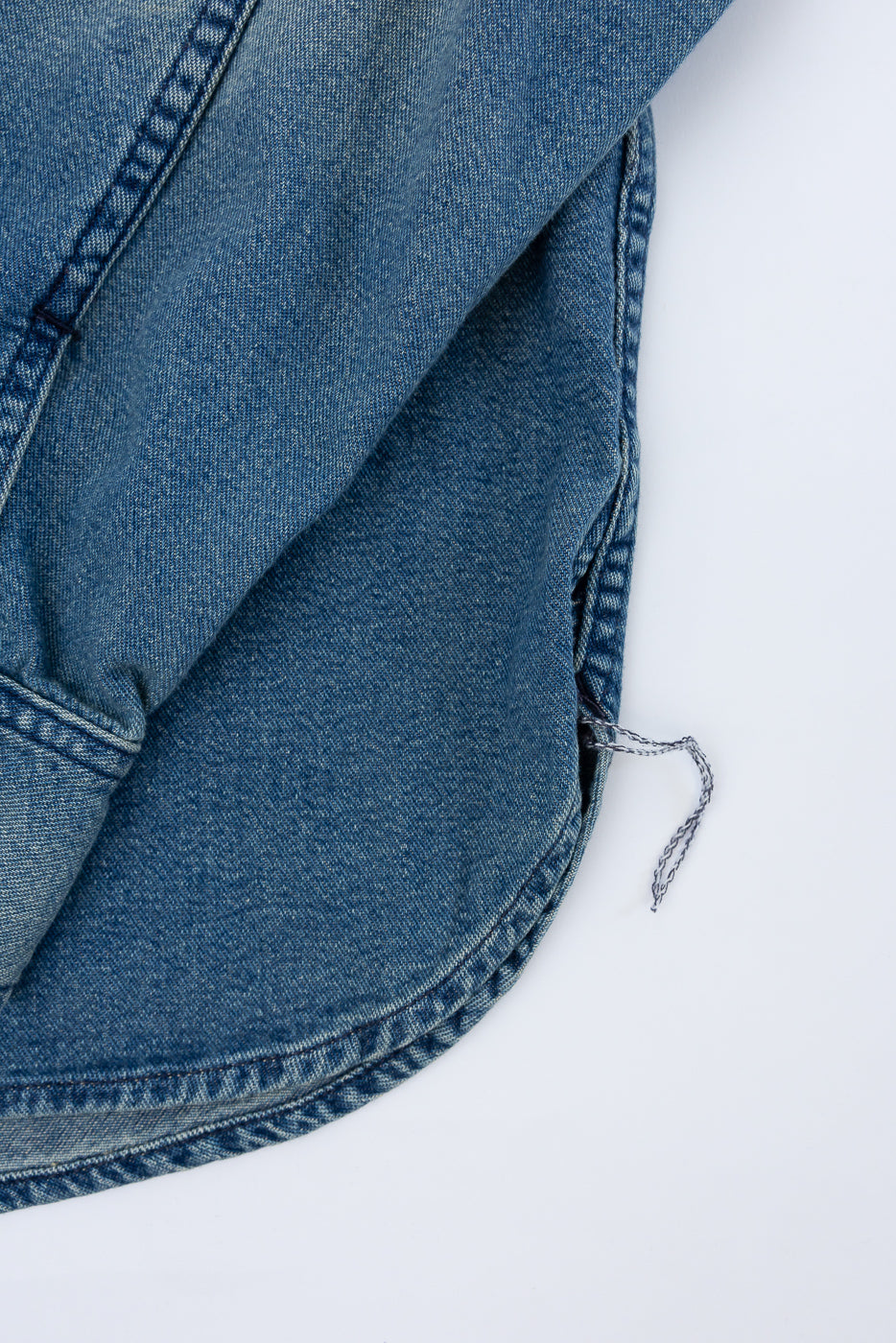 Close-up of denim shirt hem, front view, showing curved hemline, double stitching, and fabric texture.