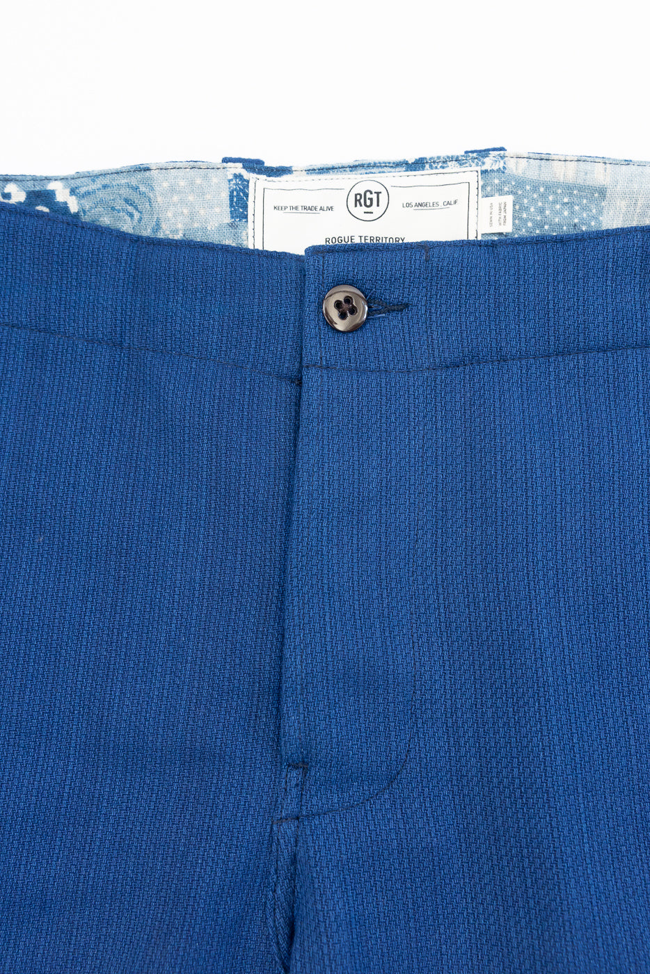 Close-up front waistband of blue men's trousers, showing button closure and RGT brand tag, textured fabric detail