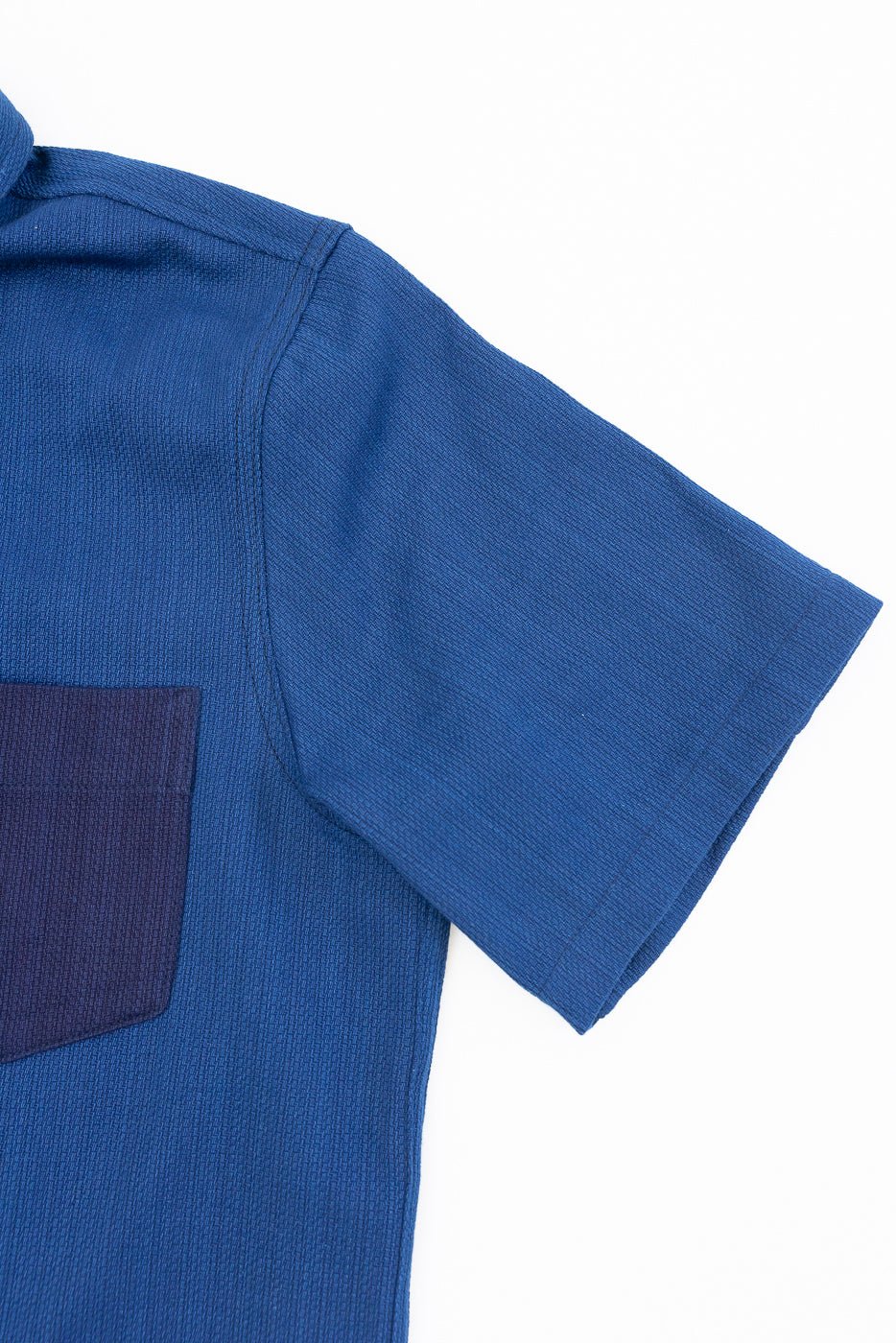 Close-up front view of blue short-sleeve shirt with ribbed texture and contrast pocket, showcasing sleeve hem and shoulder seam detail