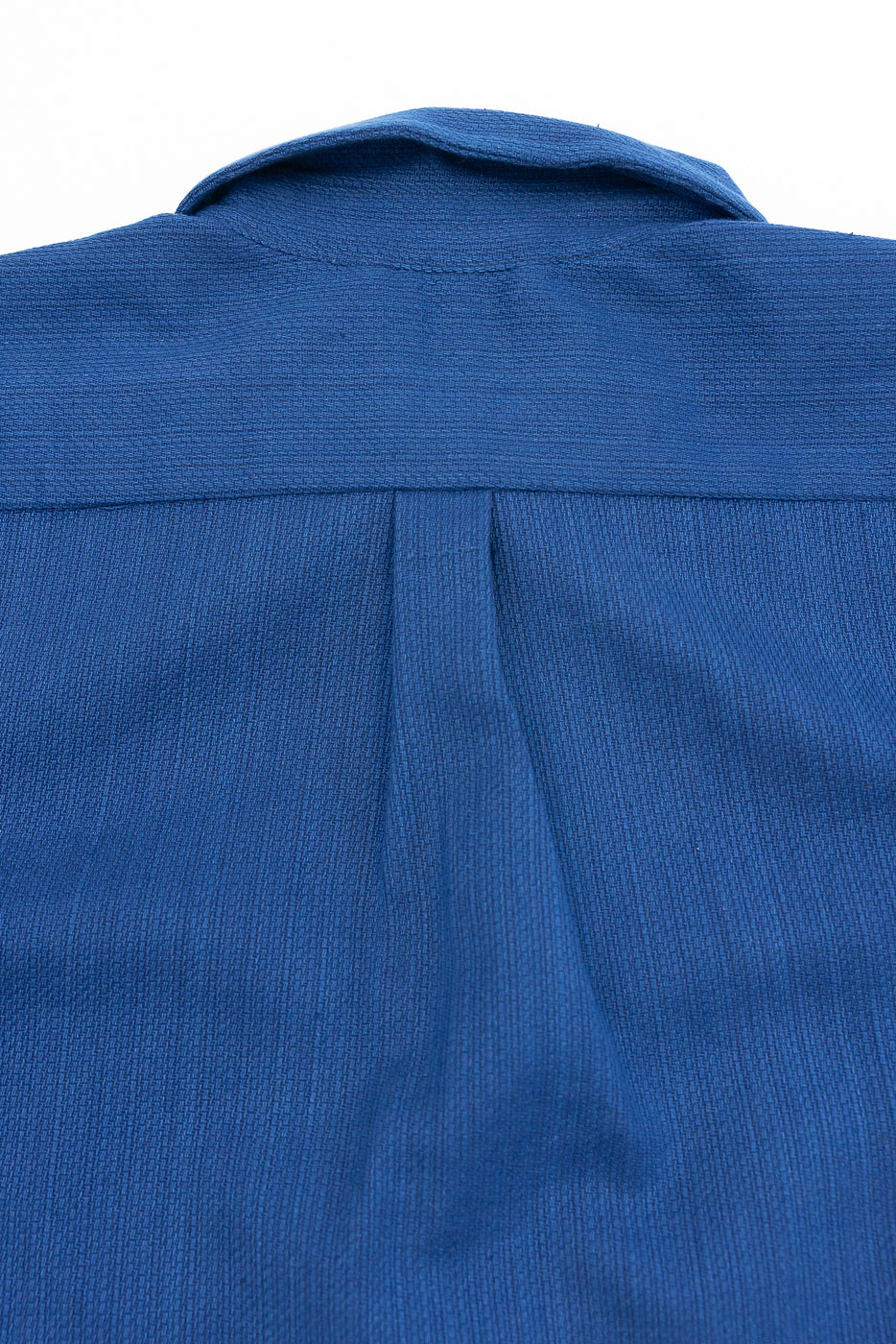 Close-up back view of blue shirt showing collar, yoke, and center pleat detail on textured fabric