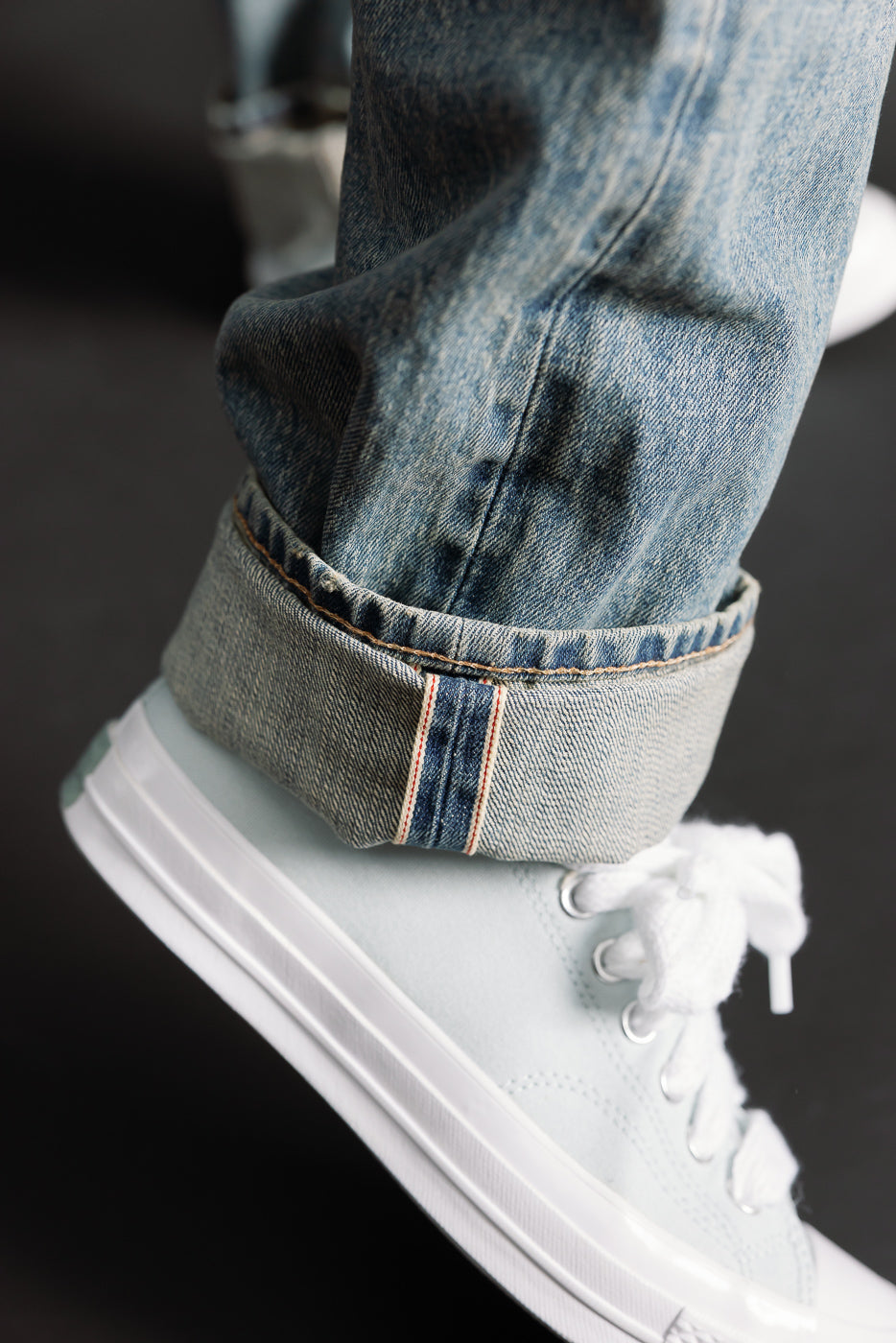 Close-up side view of men's River Washed selvedge denim jeans cuff with visible selvedge edge, worn by a male model, paired with white sneakers.