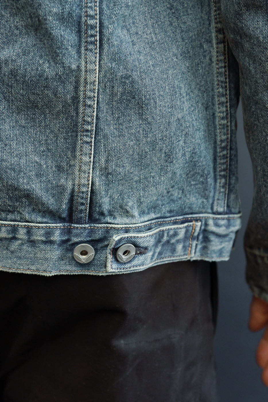 Close-up of denim jacket front, showing metal buttons and hem detail, worn by male model.