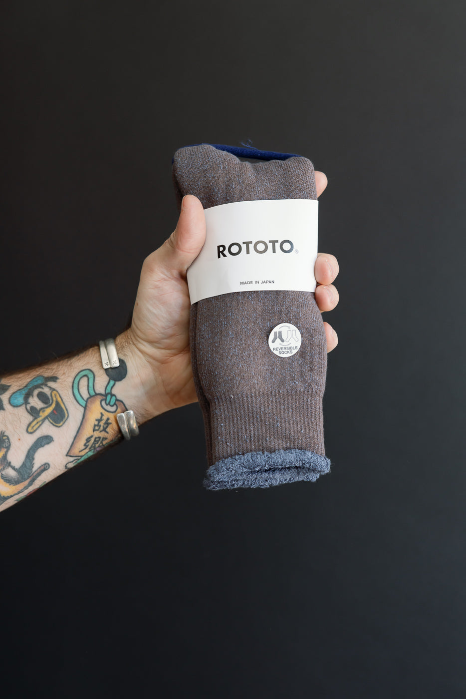 Close-up of a male hand holding Rototo reversible socks, showing fabric texture and paper tag.