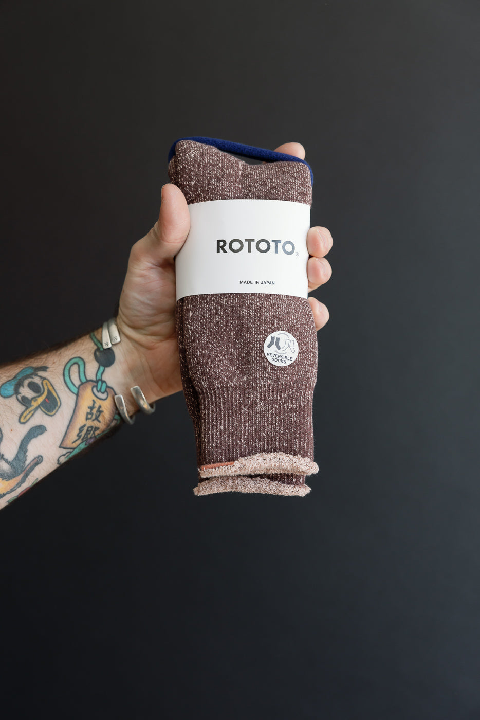 Close-up front view of a pair of brown ROTOTO reversible socks held by a male hand.