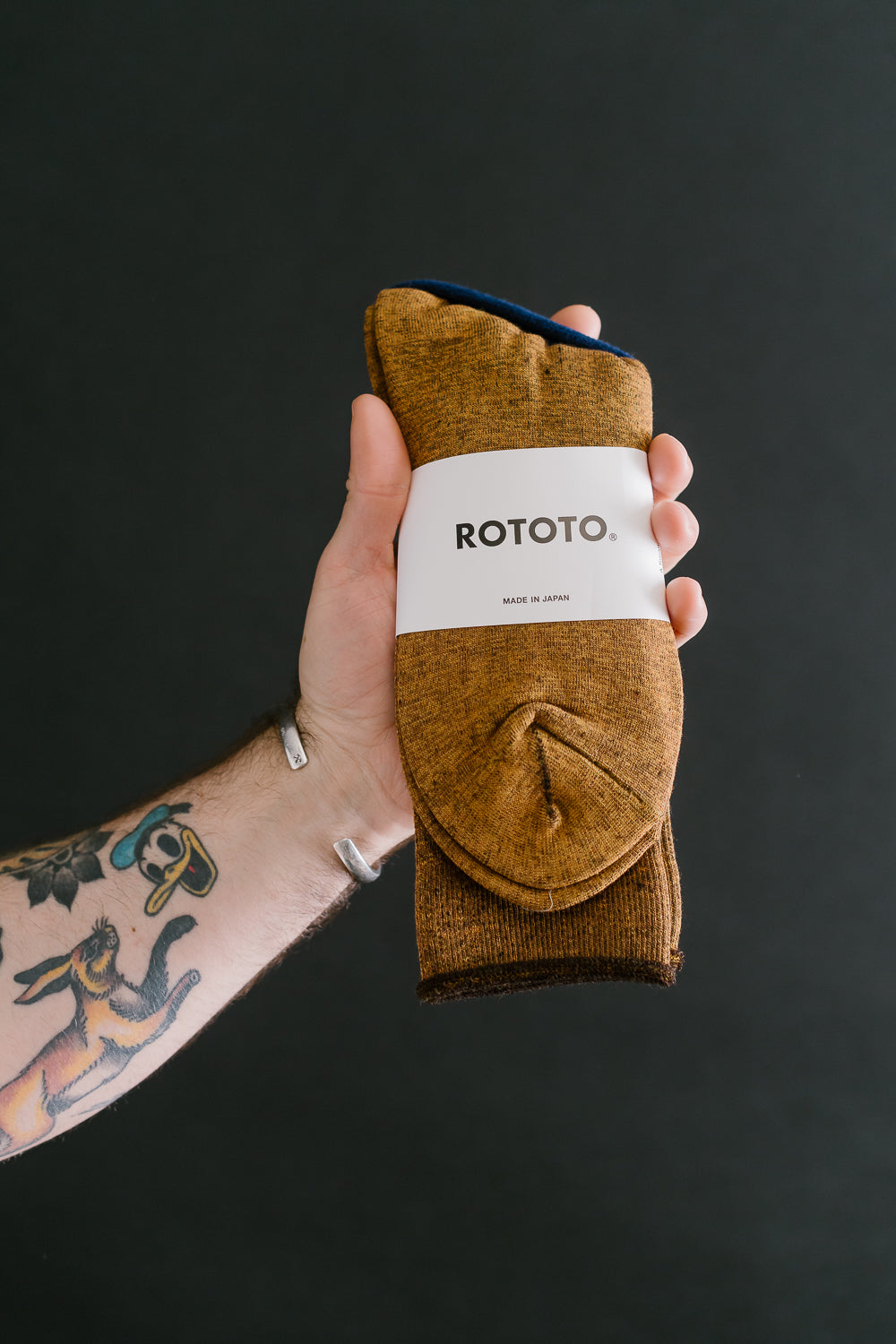 Close-up of Rototo City Crew Socks in yellow-brown, front view, held by tattooed male hand, Dant accessories