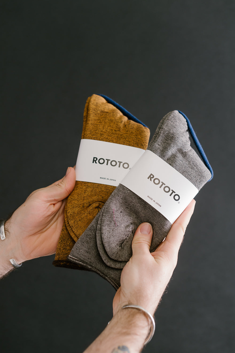 Close-up of hands holding two pairs of Rototo crew socks, front view, yellow-brown and grey colors.