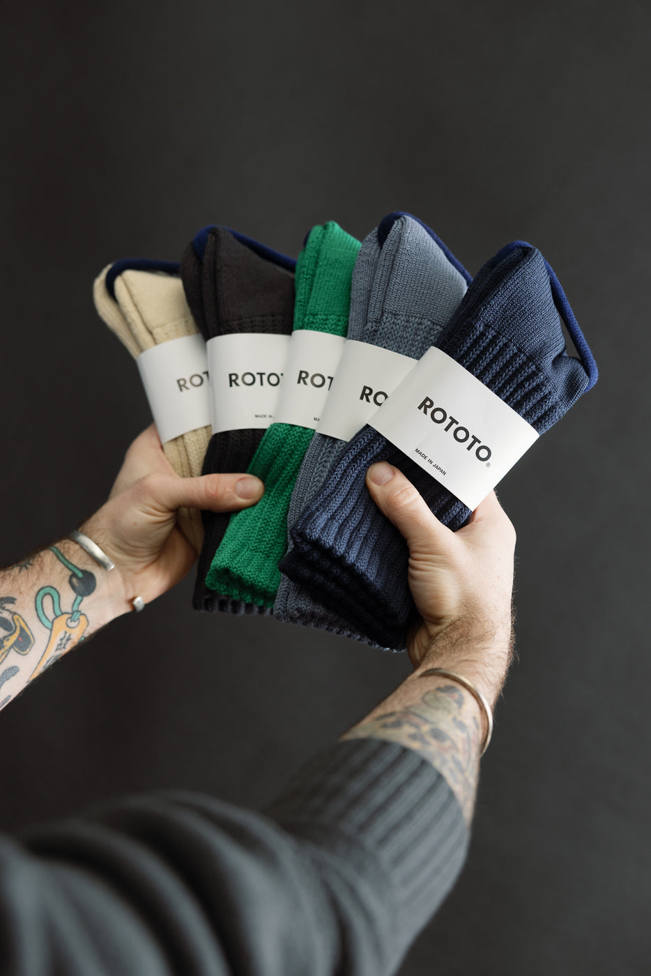 Close-up of tattooed hands holding four pairs of crew socks in beige, charcoal, green, blue; front view.