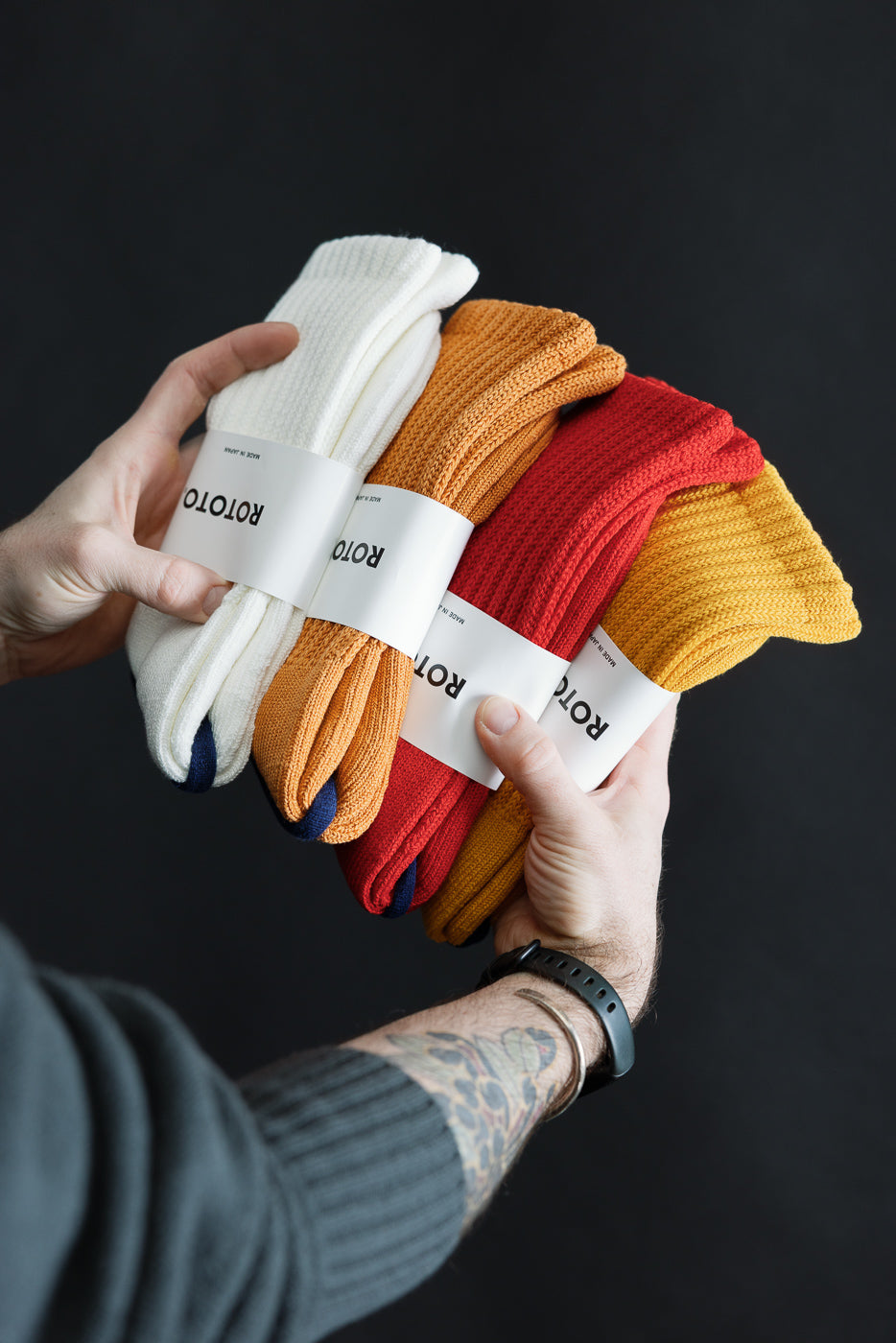 Close-up of hands holding three pairs of rototo crew socks in white, orange, and red, side view.