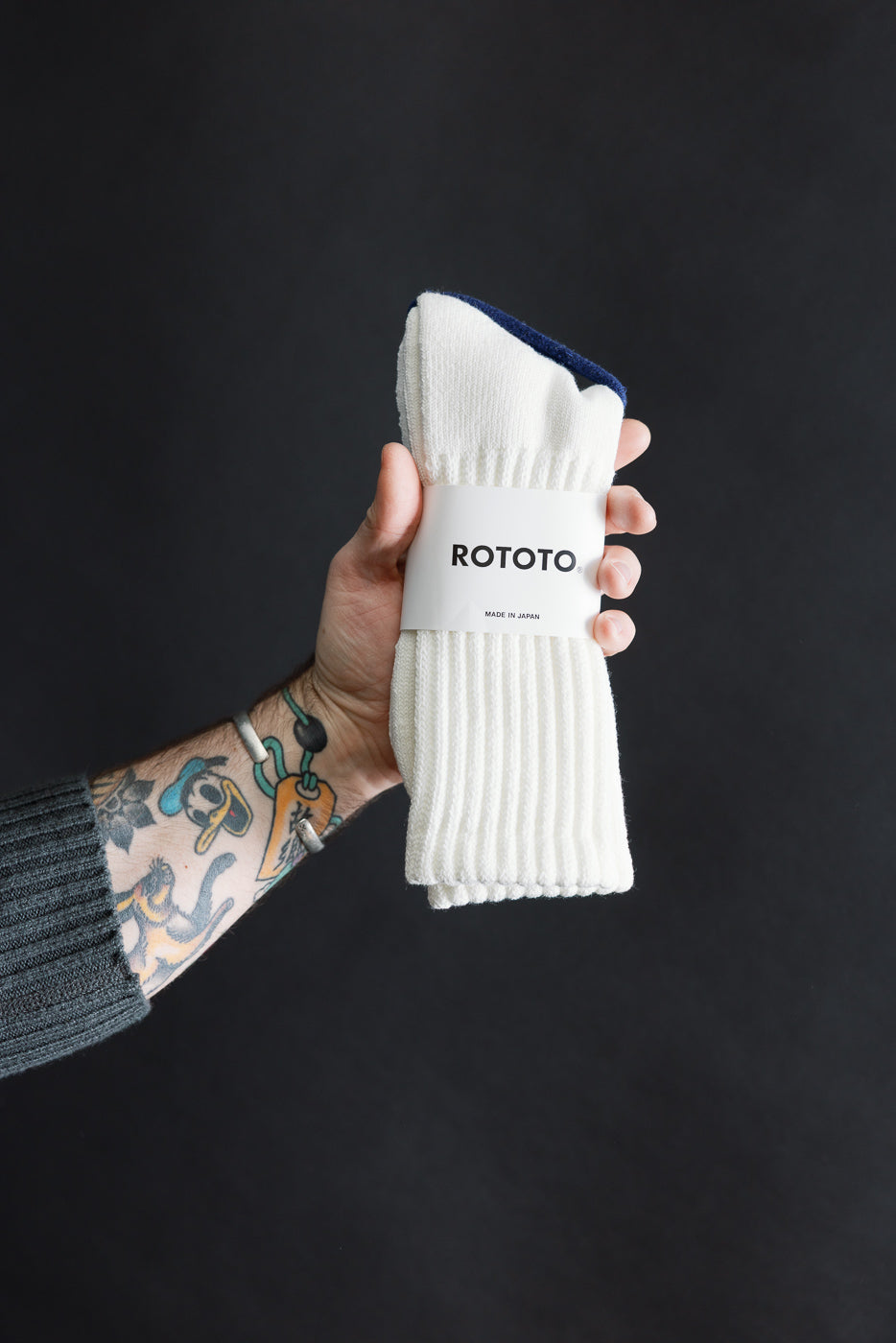 Close-up of white crew socks in packaging, front view, held by tattooed male hand