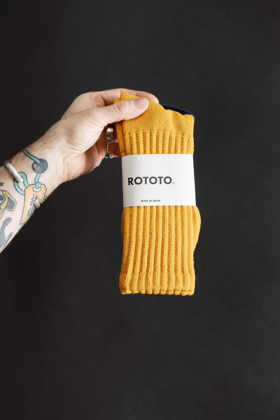 Close-up front view of yellow ribbed loose pile crew sock in packaging held by tattooed hand.