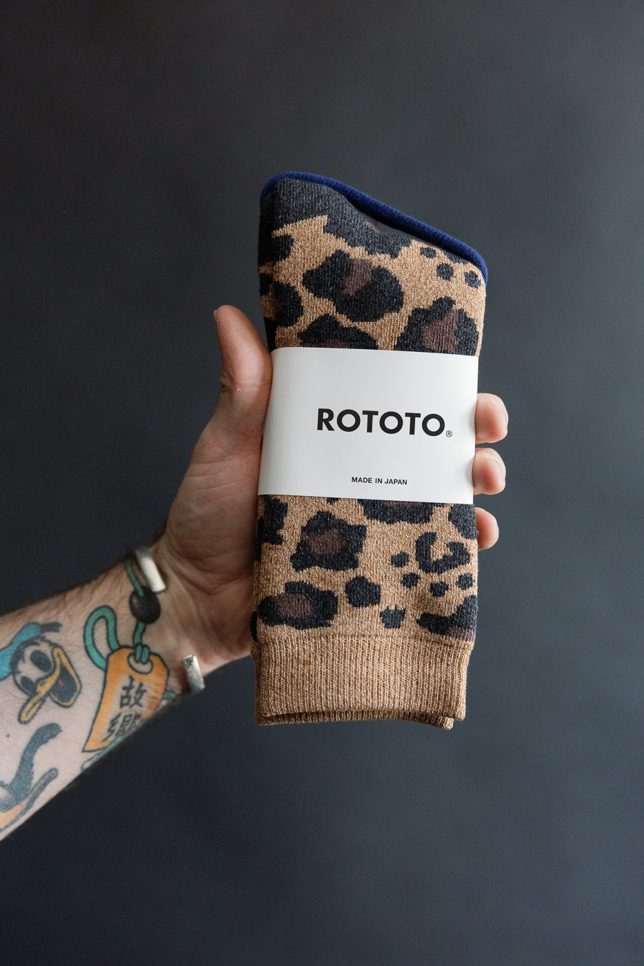 Close-up of hand holding Rototo pile leopard socks in dark beige, front of socks with label visible.