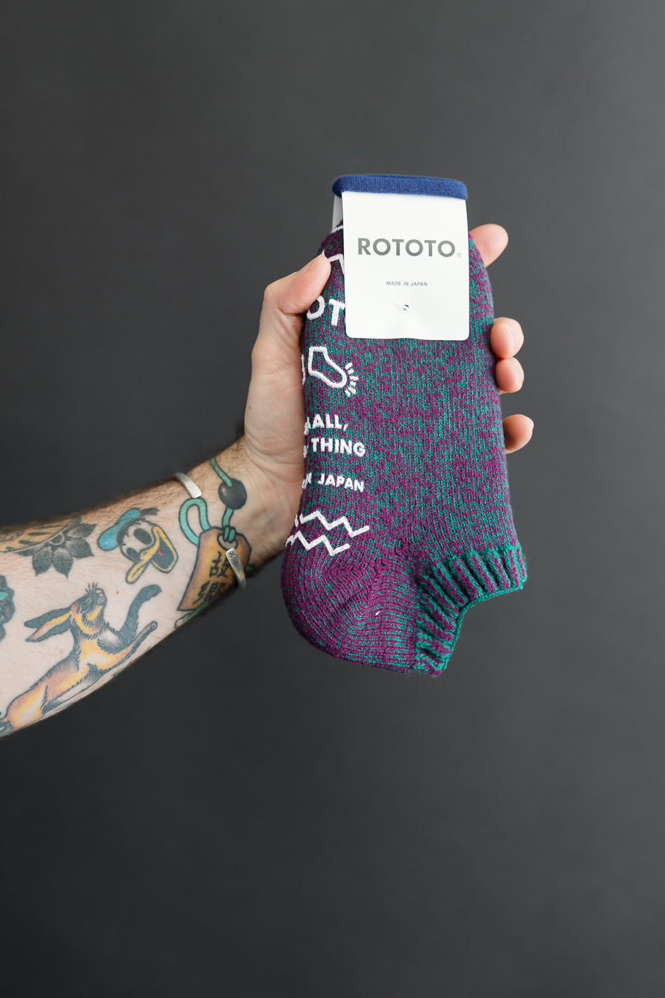 Close-up front view of patterned purple and teal knit socks held by a tattooed arm.
