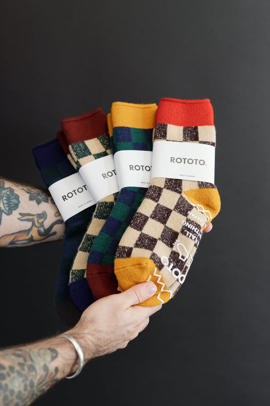 Close-up of a male's hands holding four pairs of colorful checkered Rototo socks against a dark background.