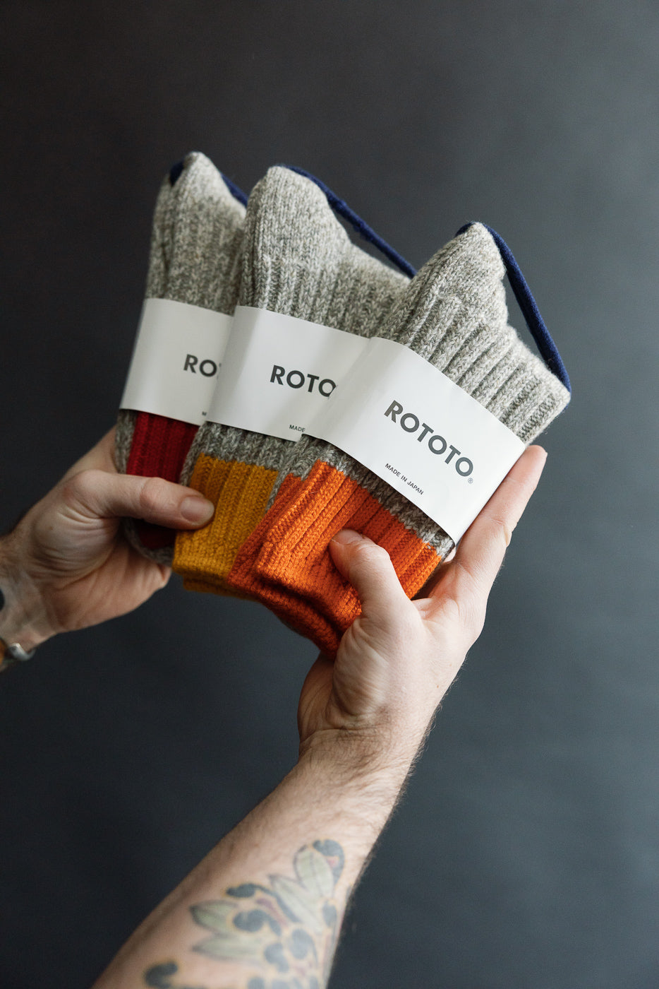 Close-up of male hand holding three pairs of ROTOTO Merino wool ribbed socks, front view, orange, yellow, and red cuffs, product detail for Dant contemporary apparel shop.