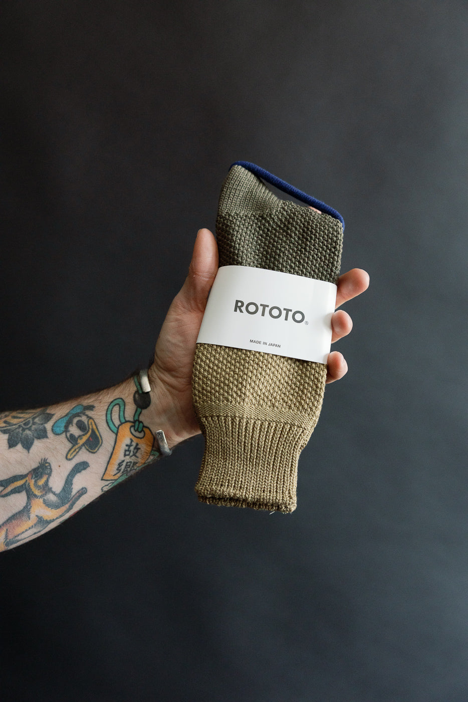 Close-up of dark olive ROTOTO seed stitch socks in packaging, front view, held in hand.