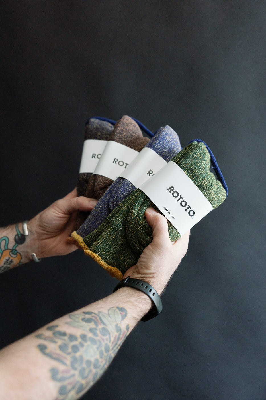 Close-up of tattooed male holding Rototo reversible pile socks, front view of folded pairs, showing Ivy Green and Dark Yellow color, contemporary casual accessory at Dant.