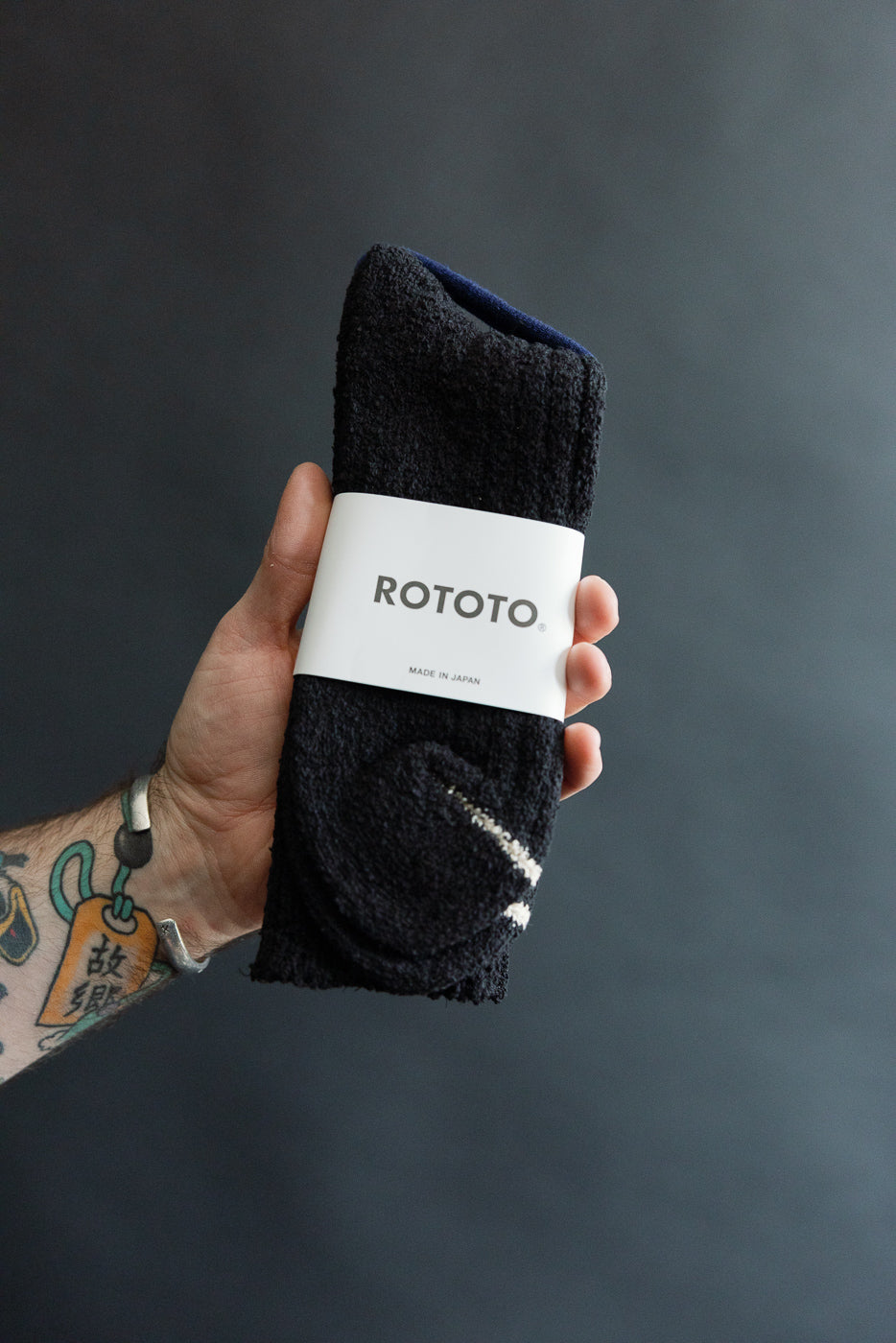 Close-up front view of black Rototo recycled cotton boucle crew socks in packaging, held in hand.
