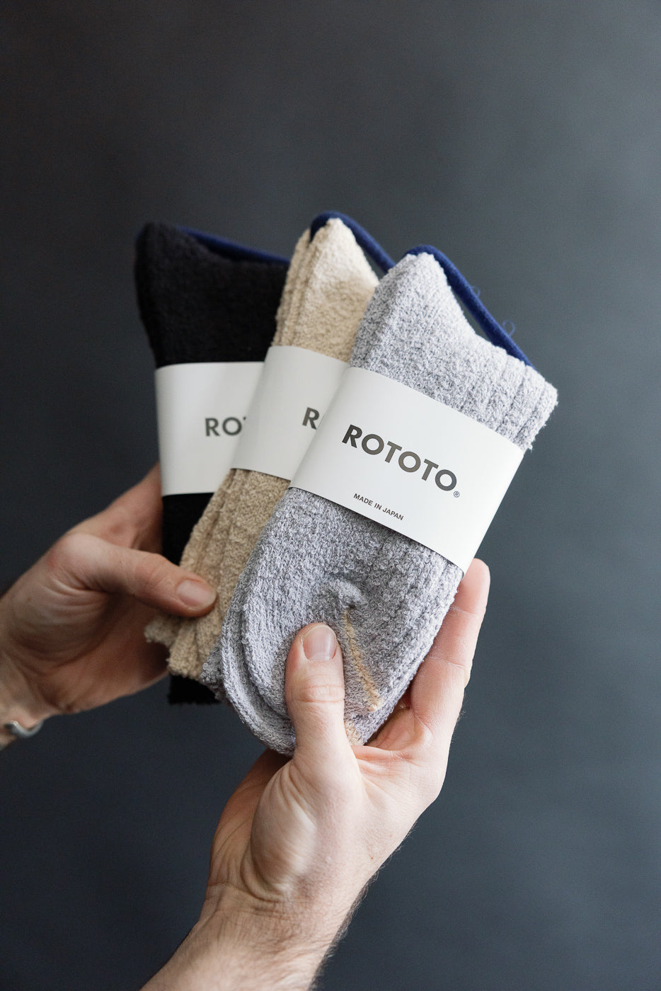 Close-up of hands holding three pairs of ROTOTO recycled cotton crew socks in black, beige, and grey, showing the front side and fabric texture for Dant contemporary casual accessories.