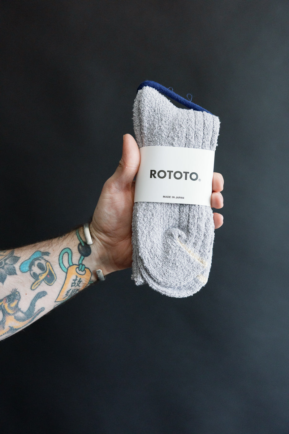 Close-up front view of Rototo recycled cotton boucle crew socks in pale blue, held by tattooed hand, showing ribbed fabric texture and brand label.