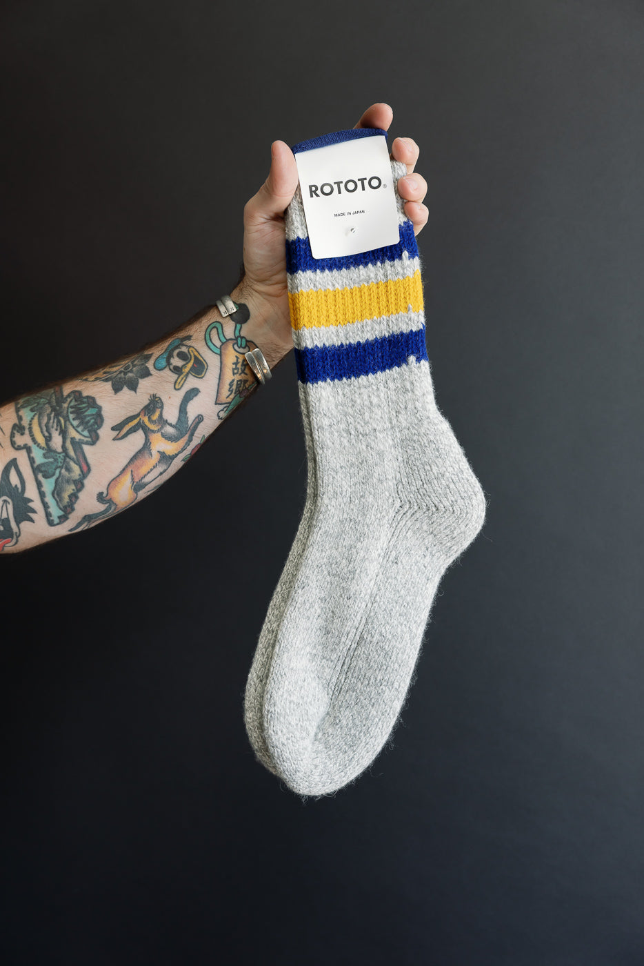Close-up front view of grey Rototo knit socks with blue and yellow stripes held by a hand.