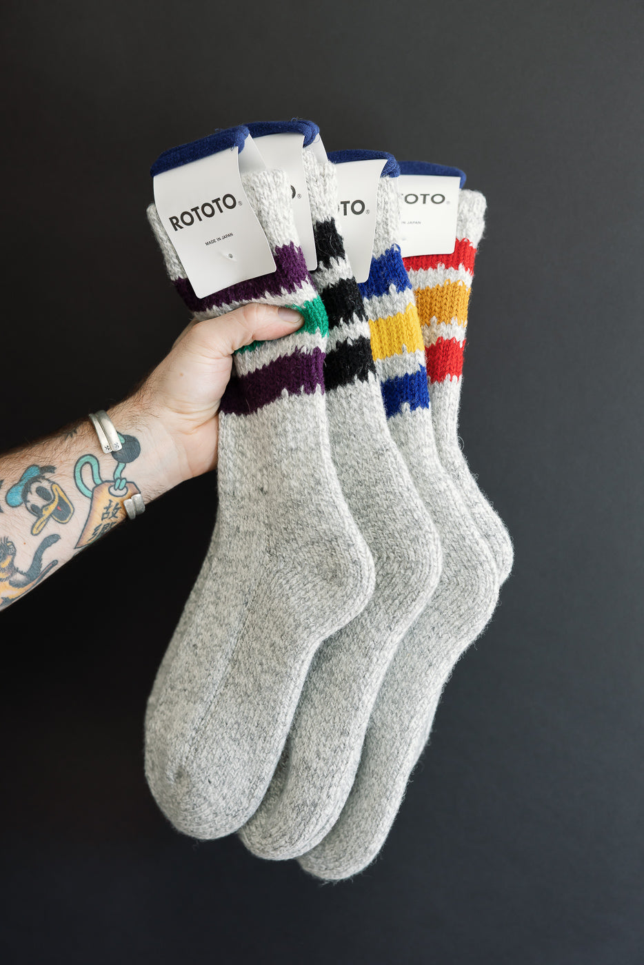 Close-up of a male hand holding three pairs of grey ROTOTO socks with colored stripes.
