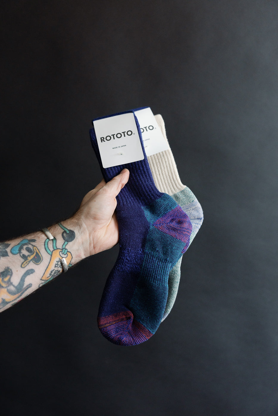 Close-up of Rototo Double Face Merino Pile Socks in dark blue, front side, held by a tattooed hand against a dark background.