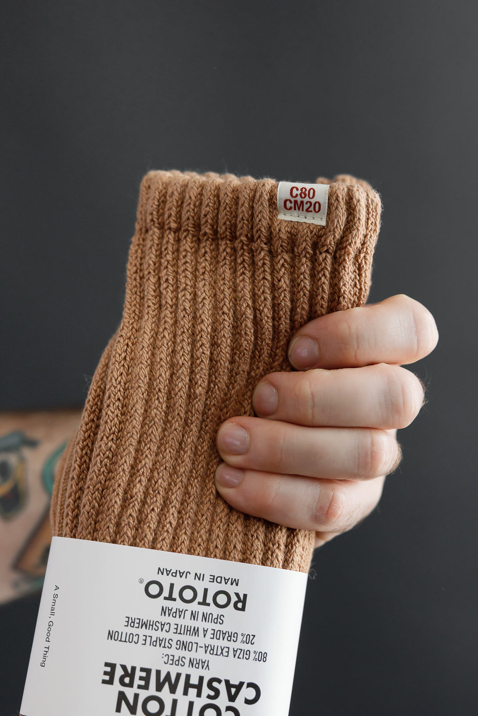 Close-up of a male model's hand holding brown ROTOTO cashmere socks, showing rib texture and tag.