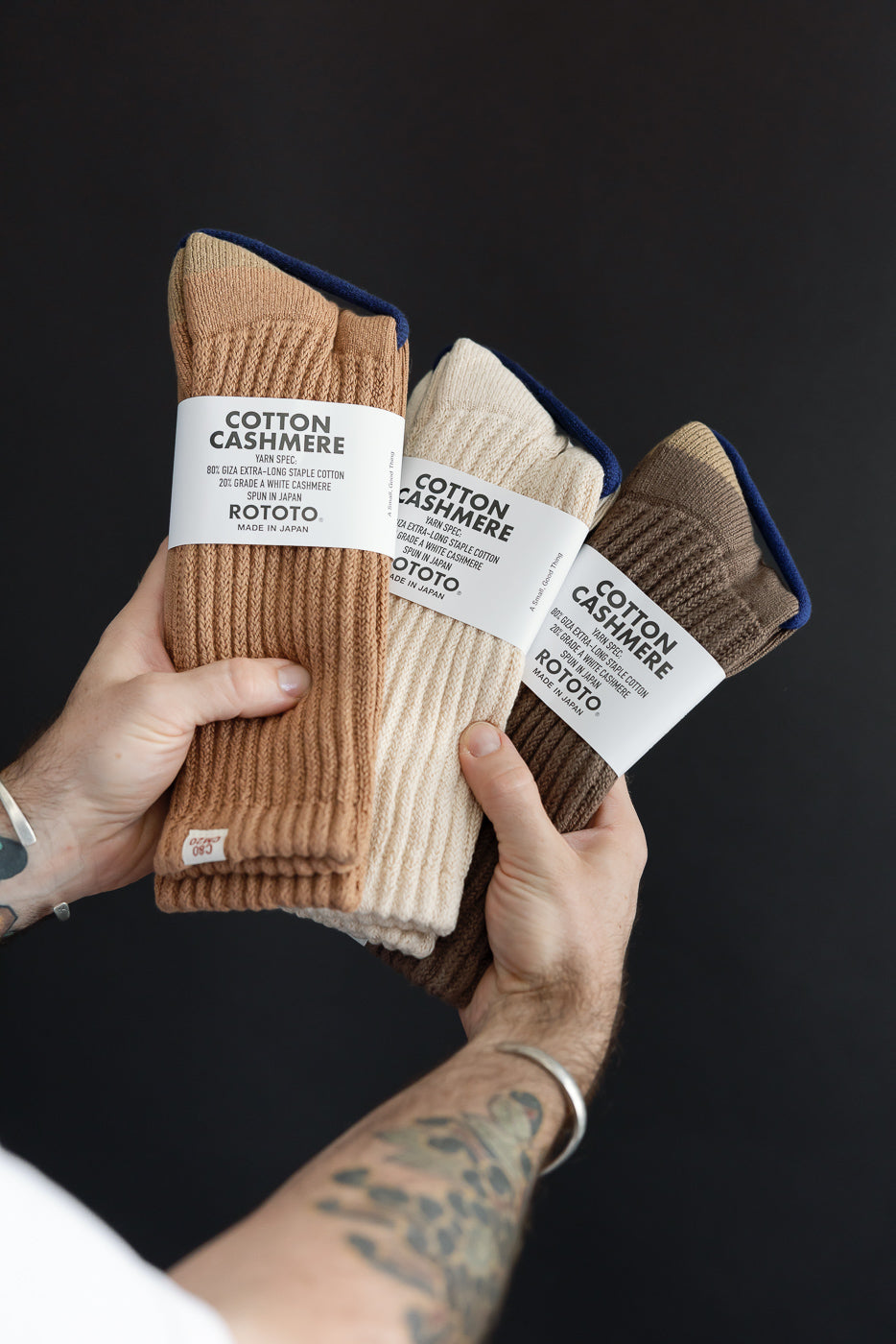 Close-up of a person's hands holding three pairs of ROTOTO cotton cashmere ribbed socks in tan, cream, and brown.