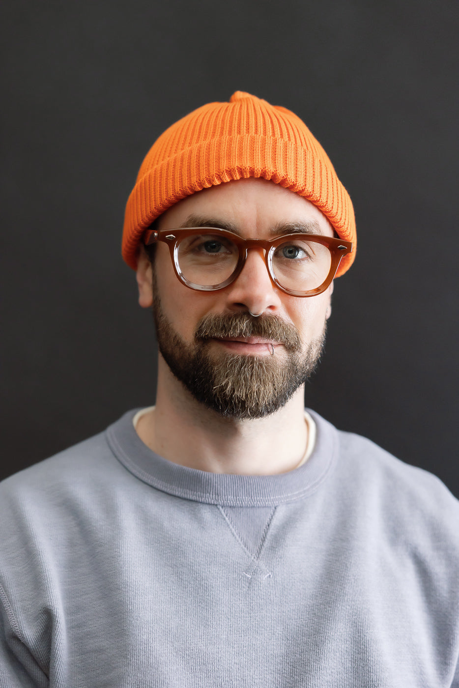 Portrait of a man wearing the R5021 - Cotton Roll-Up Beanie in bright orange, showcasing its shallow cut above the ears and 2x2 rib knit texture, styled with a light gray sweatshirt.