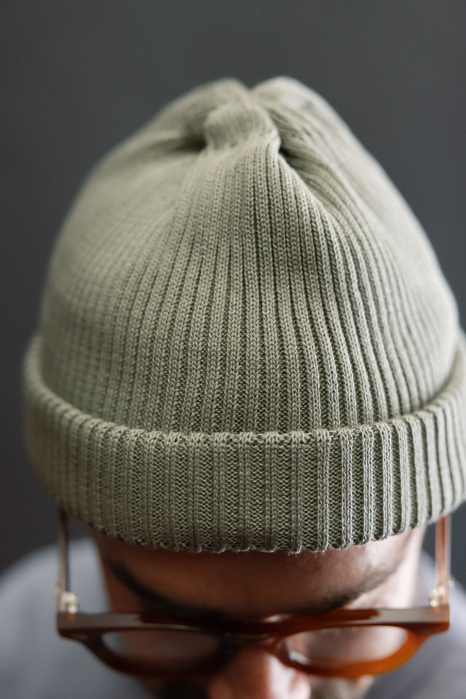 Close-up front view of sage green cotton knit roll-up beanie on male model, showing ribbed texture.