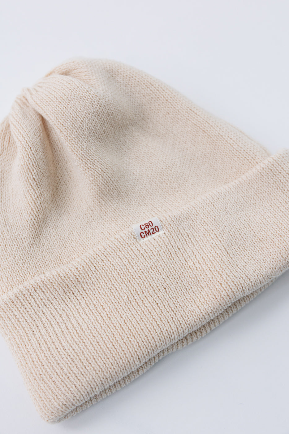 Close-up front view of a cream knit beanie featuring a small white C80 CM20 fabric tag.